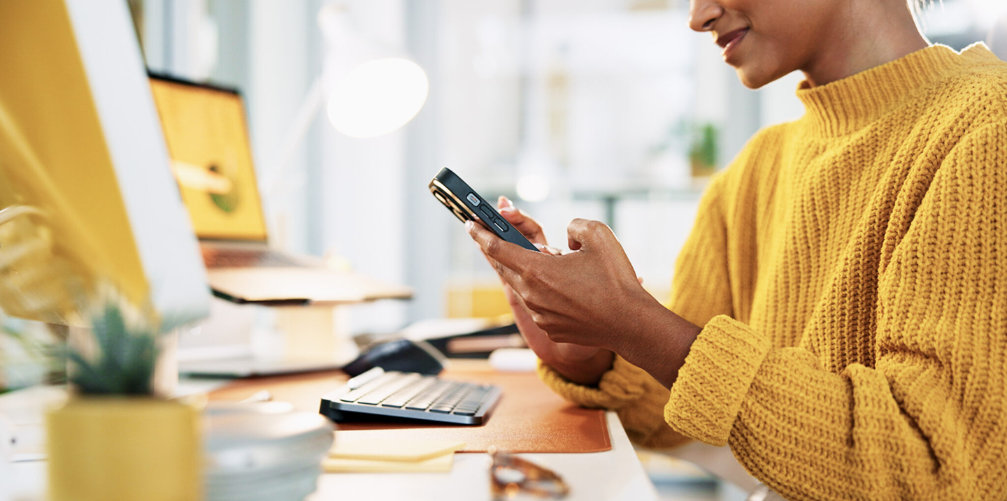 Woman in a yellow sweater using a smartphone at a desk with a computer