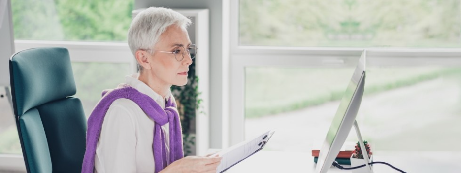 Senior woman with short gray hair and glasses working at a desktop computer in a bright, modern office
