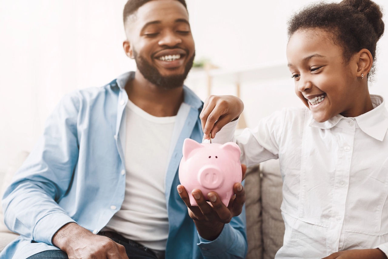 Father and young daughter smiling while she drops a coin into a pink piggy bank
