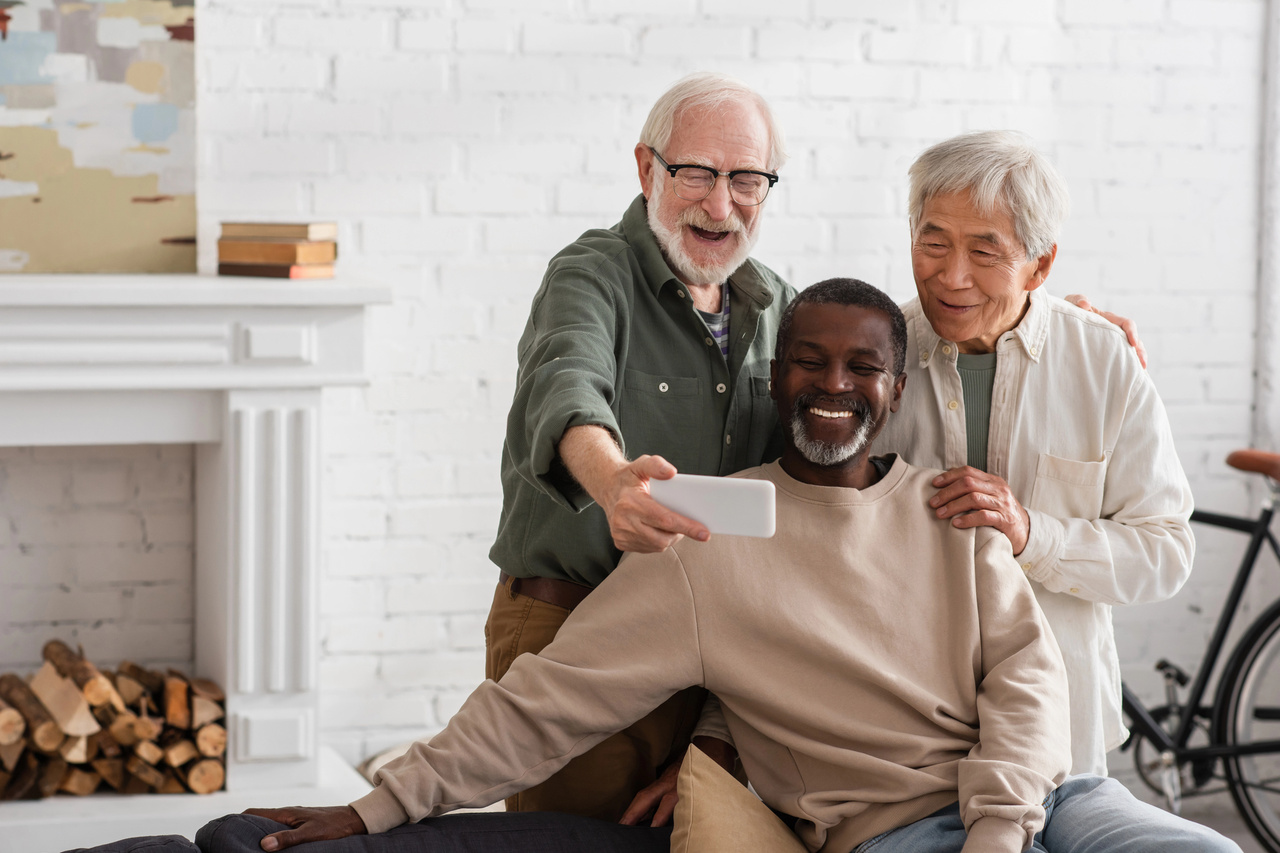 Three diverse senior men — White, Black, and Asian — laughing together while taking a selfie indoors