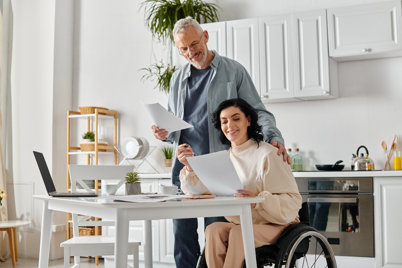 Couple reviewing financial documents at home — woman in a wheelchair smiling, husband standing beside her