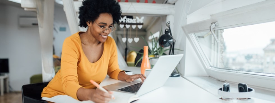 A woman in a yellow sweater smiling while working on a laptop at a bright home office desk