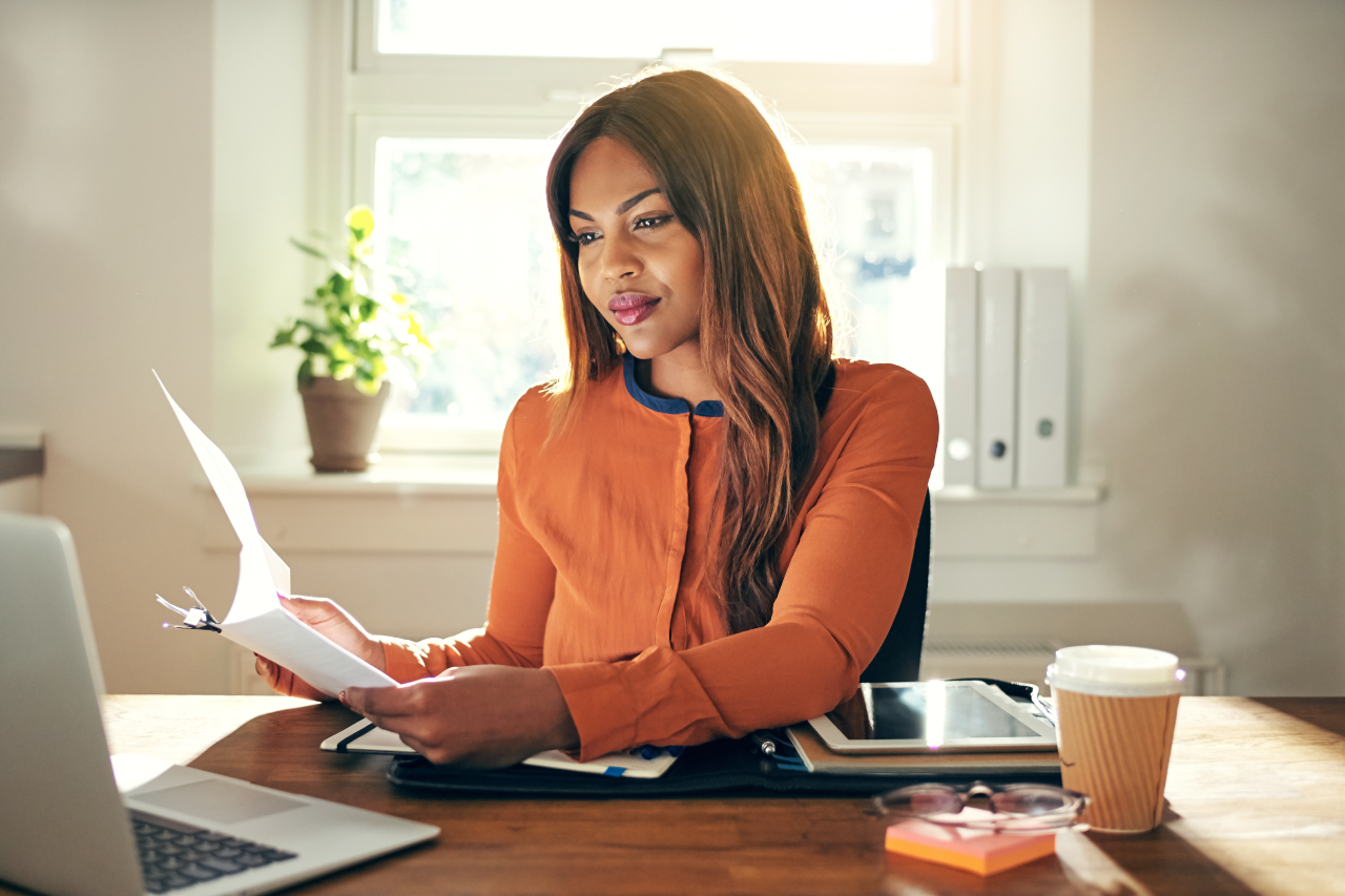 A woman in an orange top reviewing a document at her desk with a laptop and coffee nearby